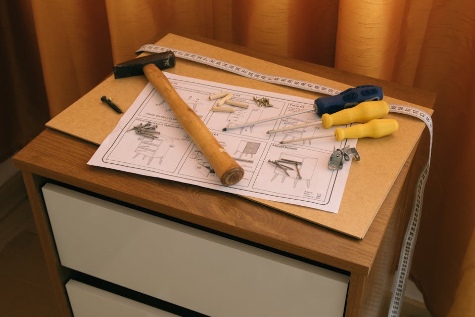 A wooden bedside table with a white drawer front is shown indoors, positioned against a background of beige curtains. On top of the table is a large, flat packing board with printed furniture assembly