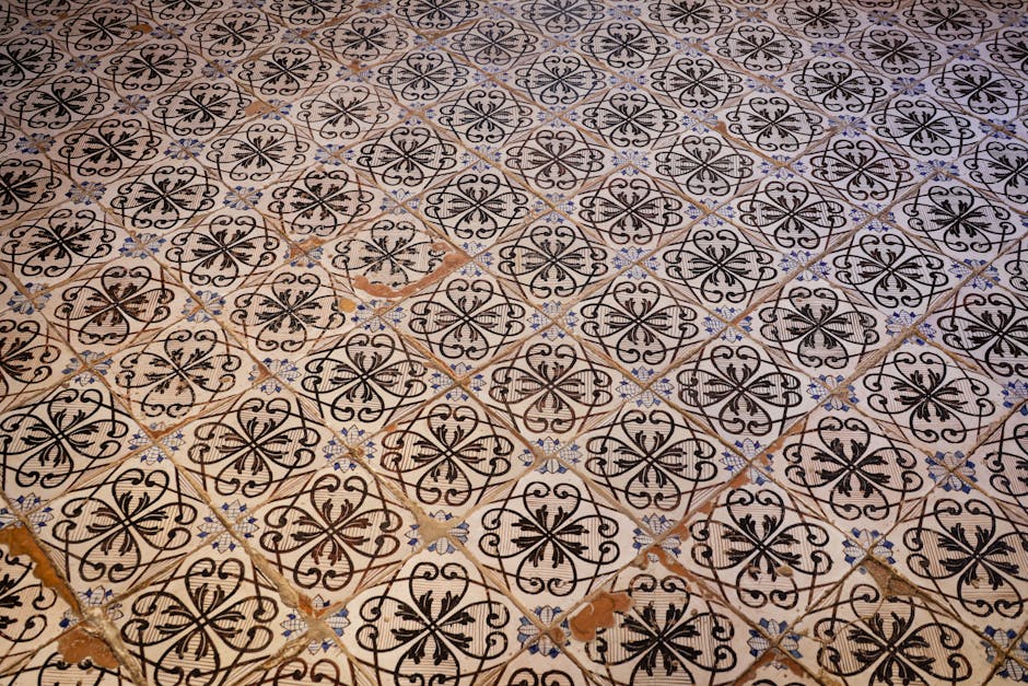 Close-up image of a patterned ceramic floor with black, white, and light blue tiles arranged in a repeating geometric design. The tiles show signs of age and wear, with some chips and discoloration vi